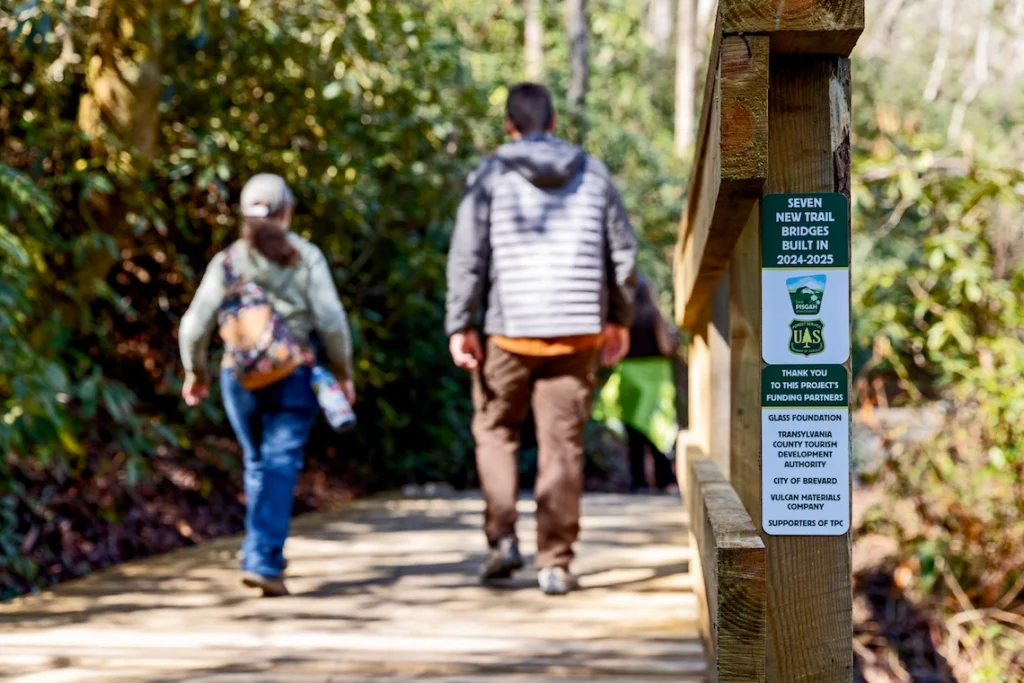 Two hikers walking on a newly constructed trail bridge on the Estatoe trail. Signs on the bridge read "seven new trail bridges built in 2024-2025" and feature partner and funder recognition.
